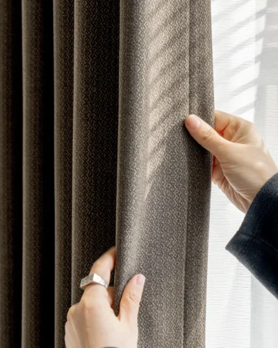 Close-up of hand touching Earthline Curtain in charcoal brown – textured fabric with soft light filtering by Lemonhone.