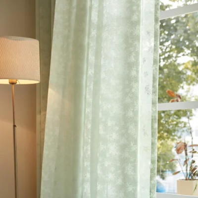 Olive Mist Curtain hanging in front of a sunlit window beside a lamp, showing soft light filtering through green botanical sheer fabric