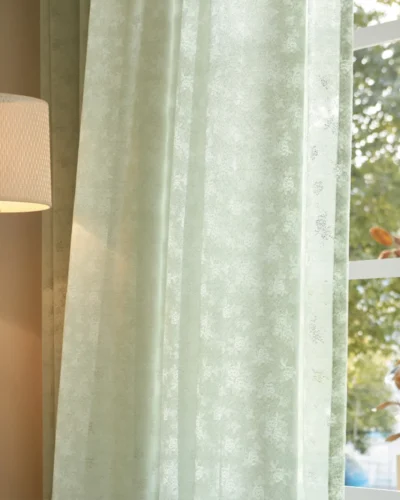 Olive Mist Curtain hanging in front of a sunlit window beside a lamp, showing soft light filtering through green botanical sheer fabric