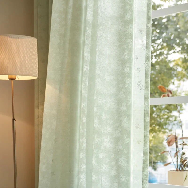 Olive Mist Curtain hanging in front of a sunlit window beside a lamp, showing soft light filtering through green botanical sheer fabric