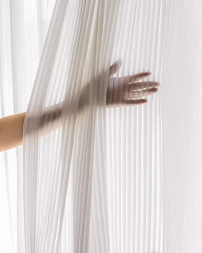 Hand behind vertical stripe sheer curtain in soft white voile, showing light-filtering privacy effect.