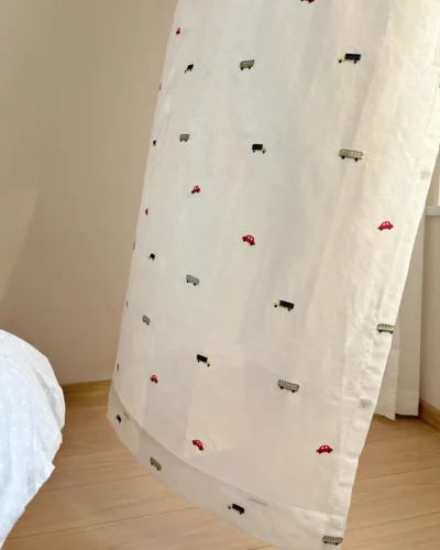 Close-up of Little Traffic sheer curtain with embroidered cars and buses hanging near the floor in a kids room.