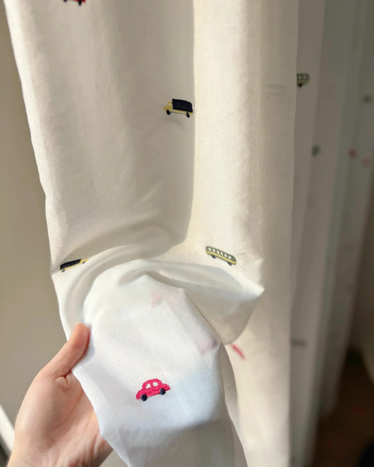 Hand holding a fabric swatch of the Little Traffic sheer curtain with embroidered red car in front of the hanging curtain.