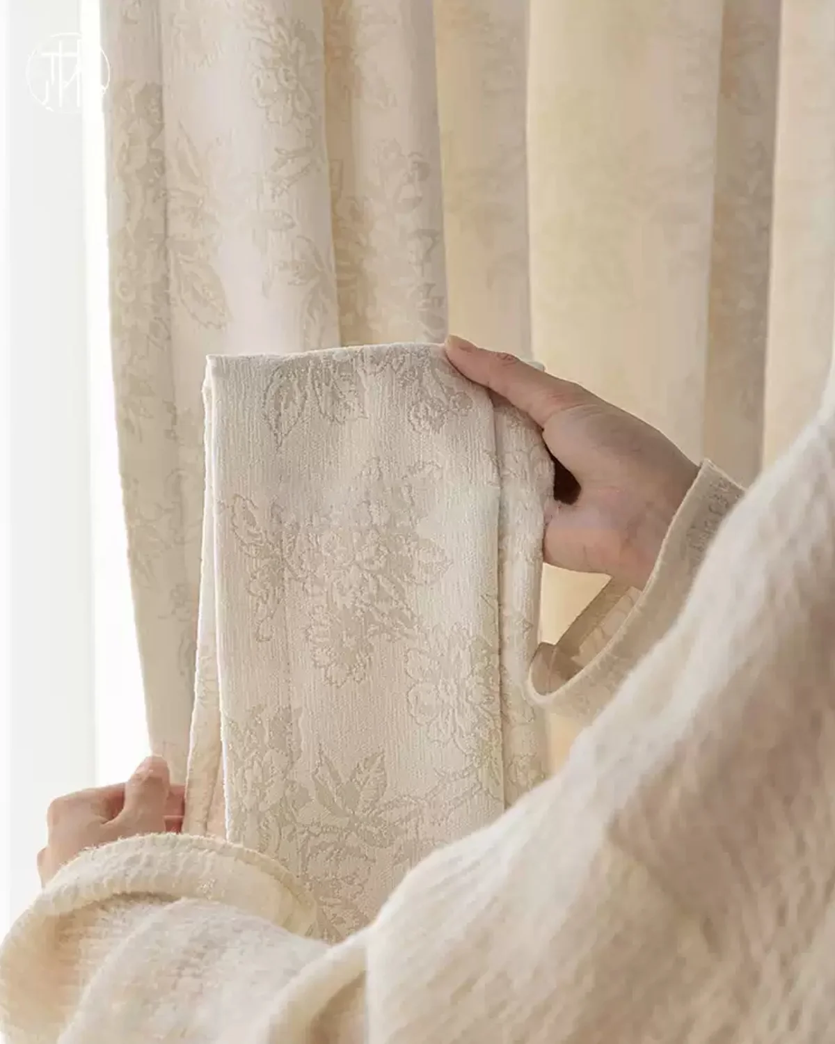 Close-up of soft beige floral jacquard curtain fabric held in hand, textured shade material.