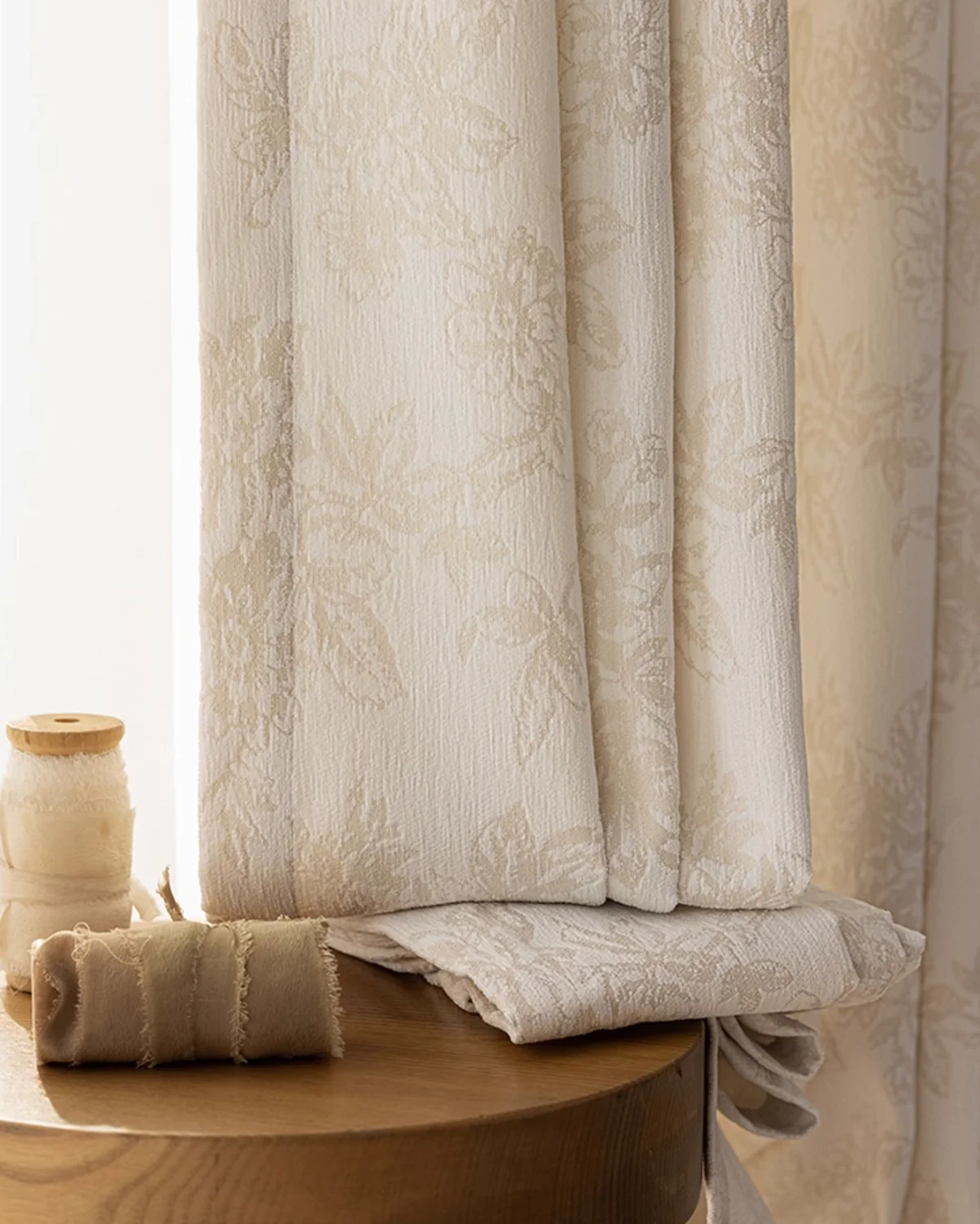 Soft beige floral jacquard curtain hanging by the window with a folded fabric sample on the table.