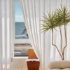 Herringbone sheer curtain in soft white textured voile hanging by a large window with ocean view and indoor plant.