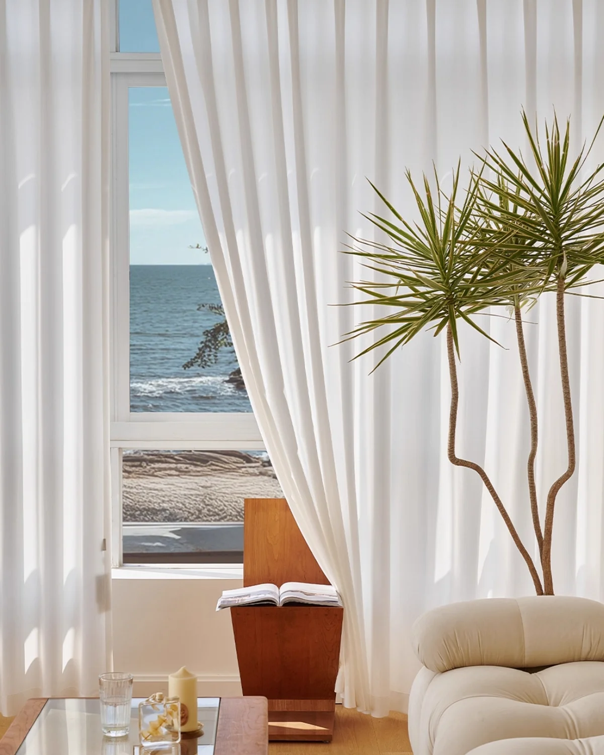Herringbone sheer curtain in soft white textured voile hanging by a large window with ocean view and indoor plant.
