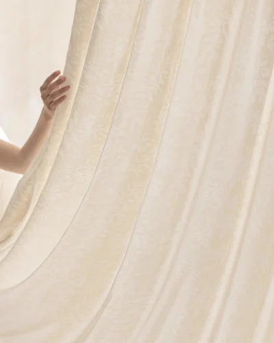 Hand lifting the Blossom Whisper sheer curtain to show its soft drape and light-filtering beige floral jacquard fabric