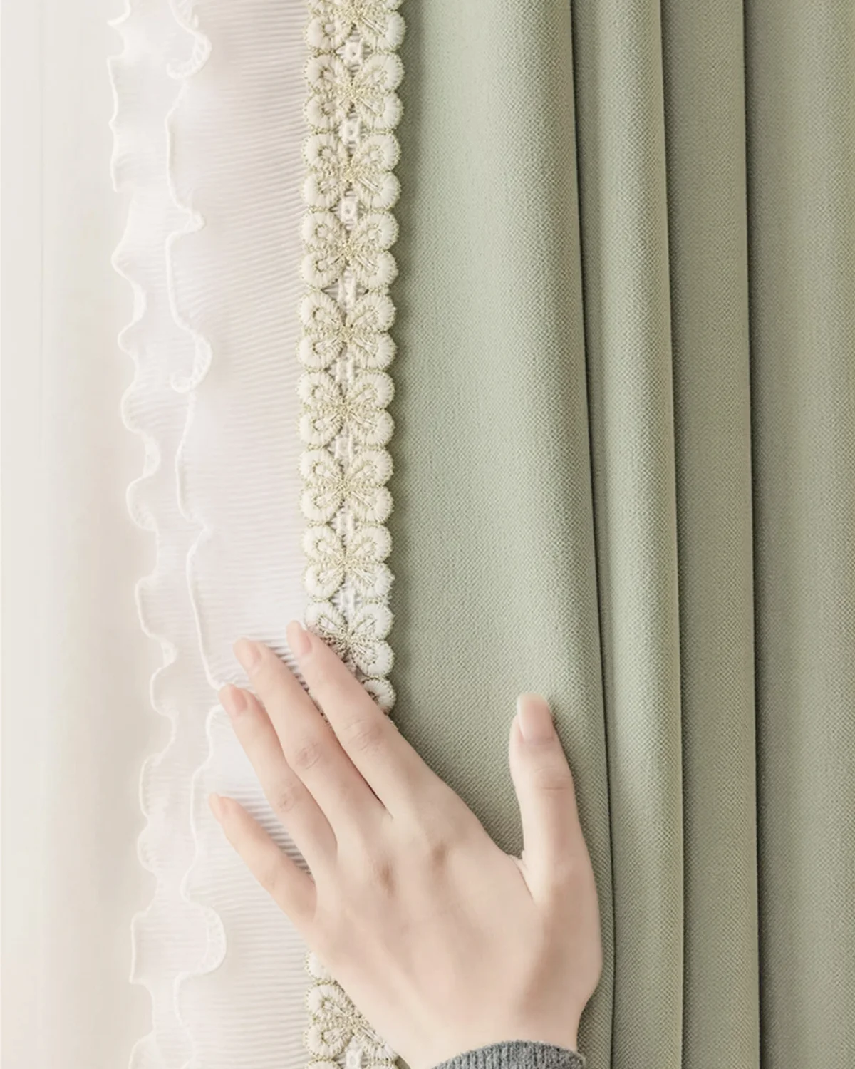 Hand touching white sheer curtain layered beside a sage green drape, showing texture and contrast