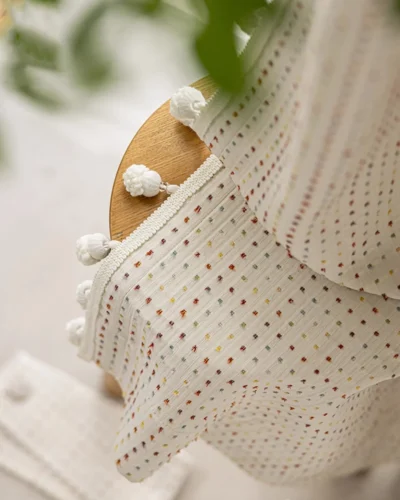 Close-up of the Confetti Pom Pom Curtain with multicolour dotted weave and white pom pom trim draped over a wooden stool.