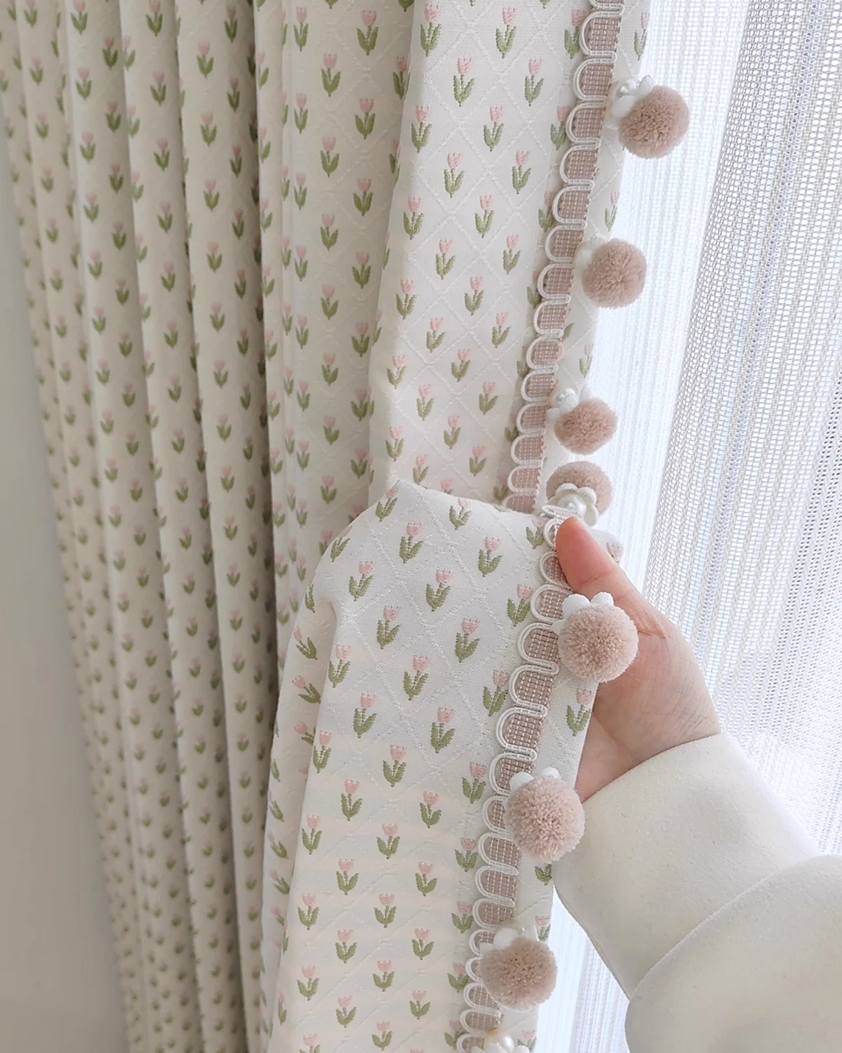 Hand holding embroidered sheer curtain showing petite floral pattern and pom pom trim edge