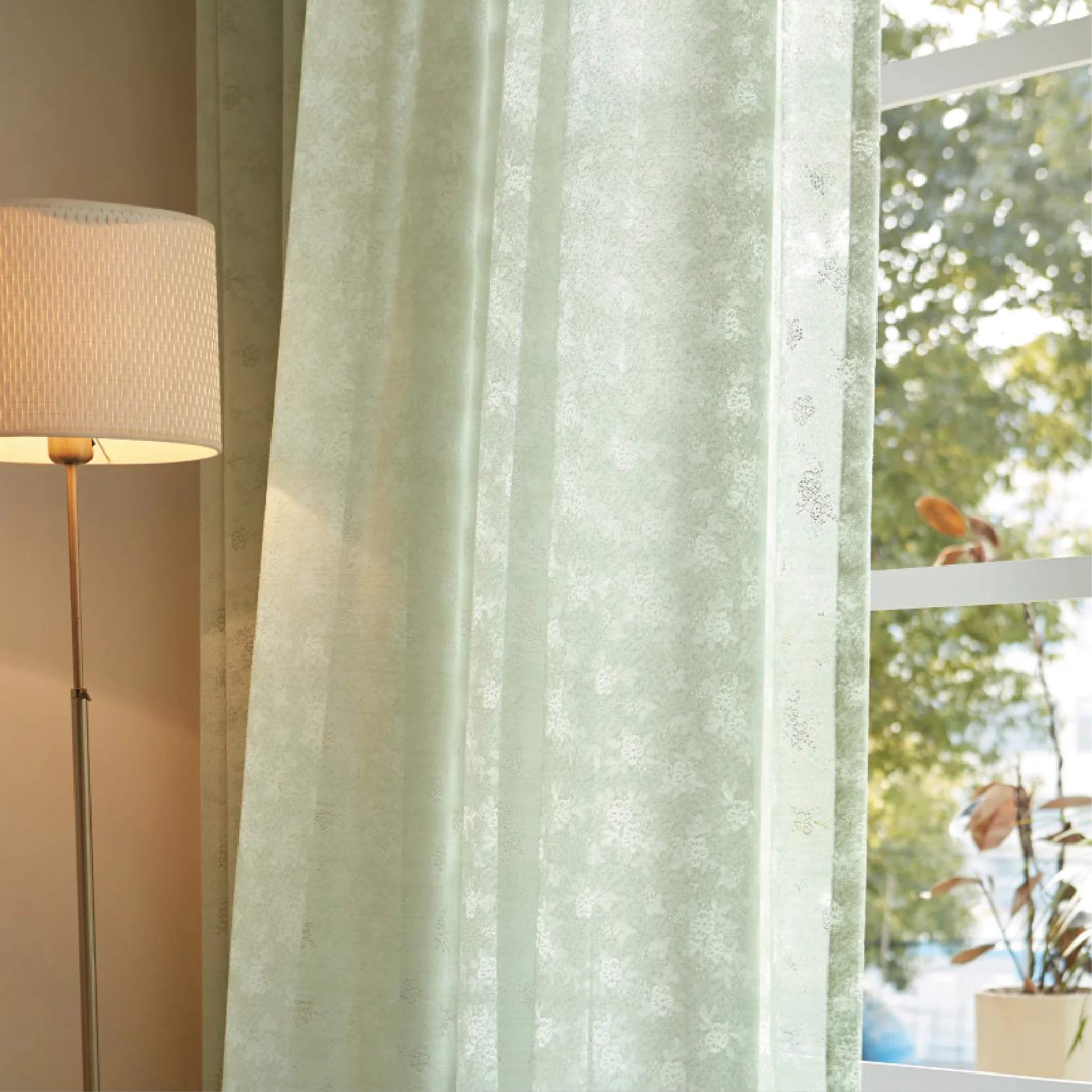 Olive Mist Curtain hanging in front of a sunlit window beside a lamp, showing soft light filtering through green botanical sheer fabric