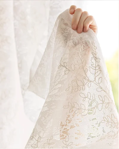 Hand holding rope-embroidered floral sheer curtain in ivory—close-up of light-filtering lace texture.