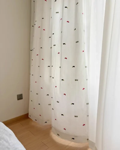 Little Traffic sheer curtain with embroidered cars and buses hanging next to the bed in a kids room.