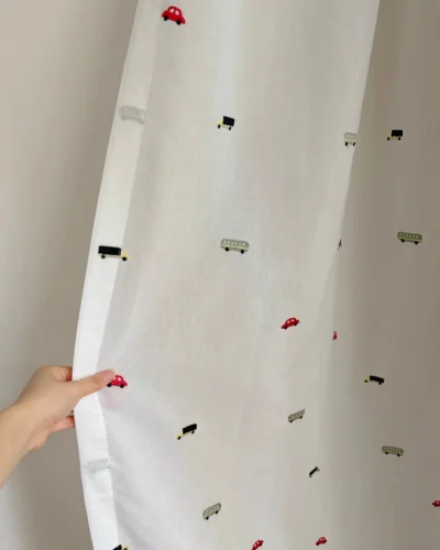Hand holding the Little Traffic sheer curtain with embroidered cars and buses to show the overall pattern and fabric transparency.