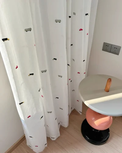 Little Traffic sheer curtain with embroidered cars and buses hanging beside a small round table in a kids room.