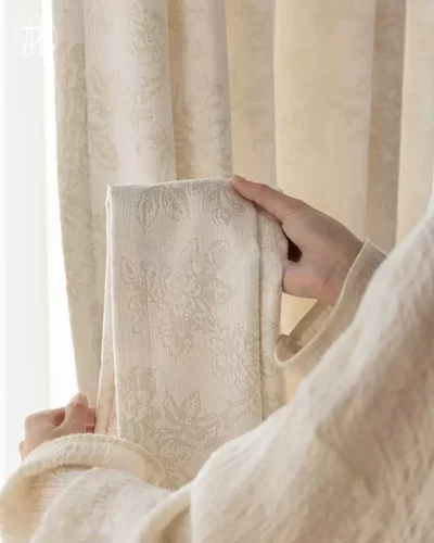 Close-up of soft beige floral jacquard curtain fabric held in hand, textured shade material.