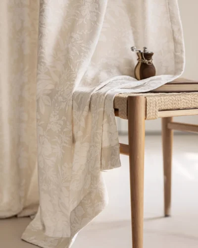Blossom Whisper sheer curtain in beige floral jacquard pooling softly on the floor beside a wooden side table with a small vase