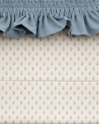 Close-up Roman shade in off-white with small blue tulip pattern and green leaves, topped with a soft blue ruffle valance.