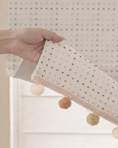 Hand holding the Rainbow Sprinkles Roman Shade hem showing fabric texture and pom-pom trim