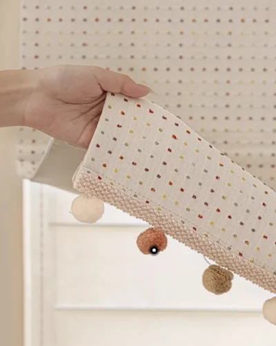 Hand holding the Rainbow Sprinkles Roman Shade hem showing fabric texture and pom-pom trim