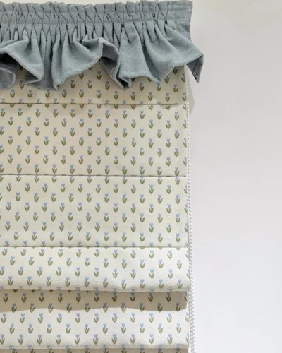 Close-up Roman shade in off-white with small blue tulip pattern and green leaves, topped with a soft blue ruffle valance.