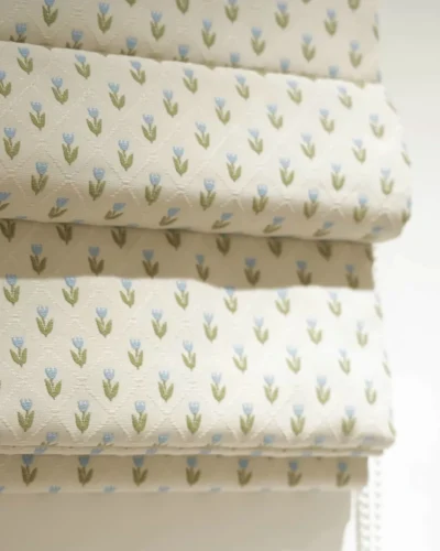 Roman shade close-up in off-white with small blue tulip pattern and green leaves, showing layered folds and soft texture.