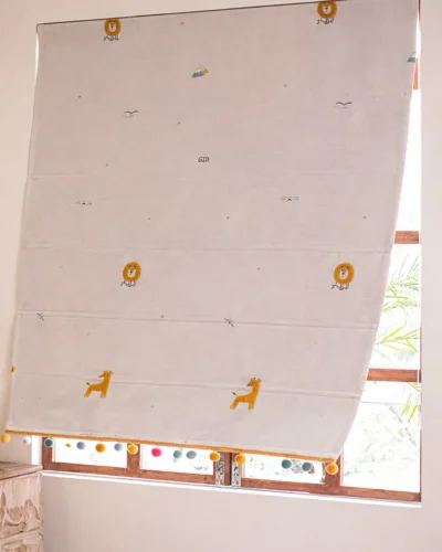 Safari embroidered Roman shade with colorful pom-pom trim, shown half-lowered on a wooden window.