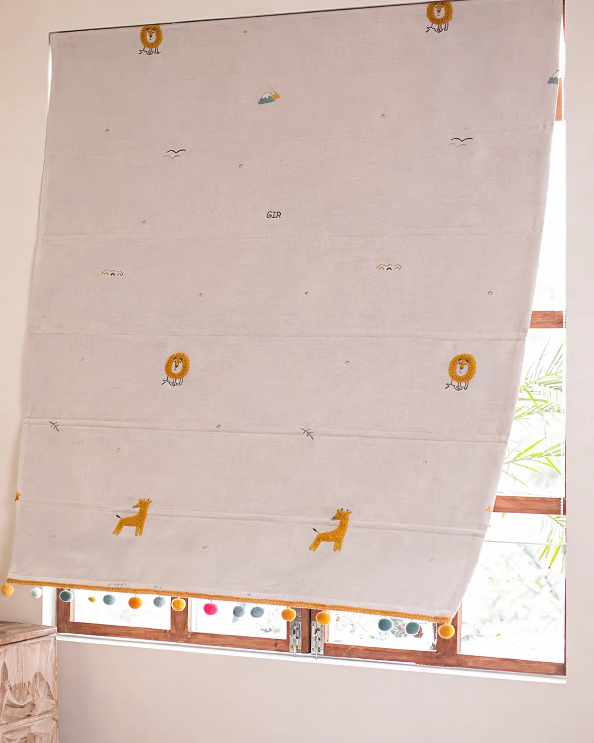 Safari embroidered Roman shade with colorful pom-pom trim, shown half-lowered on a wooden window.