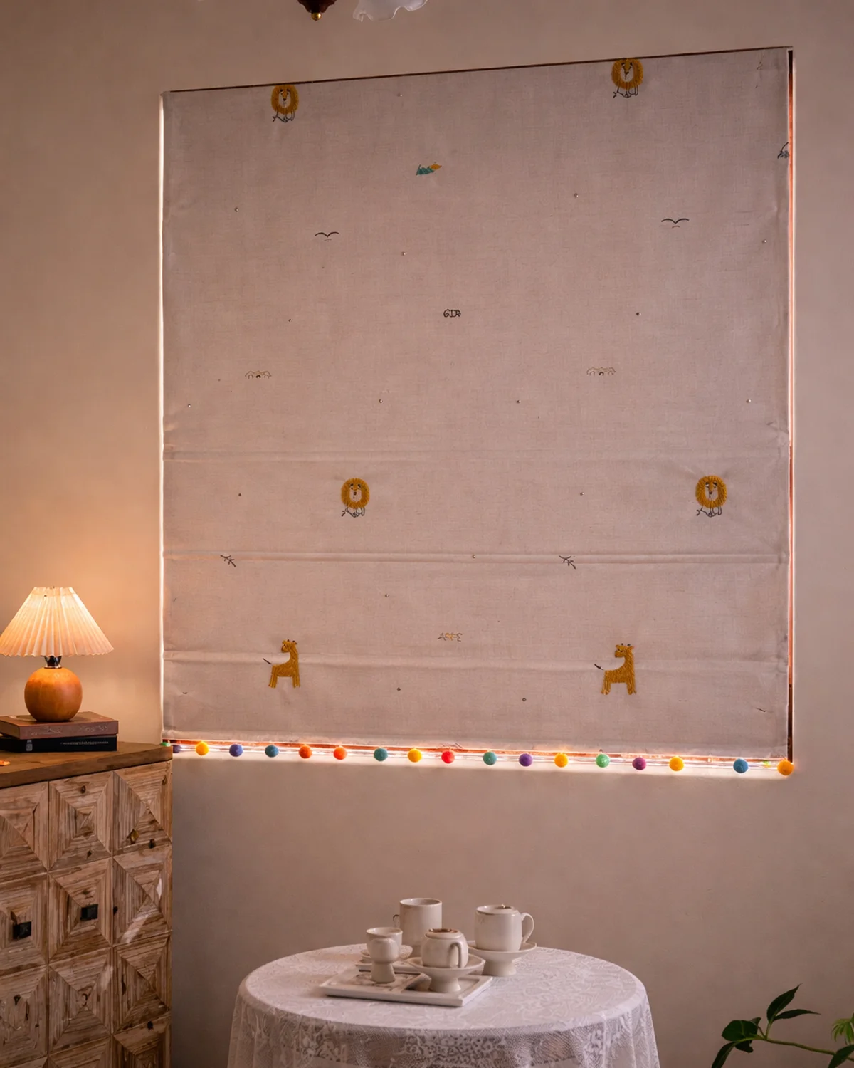 Whimsical safari embroidered Roman shade with colorful pom-pom trim, styled in a cozy kids room at night.
