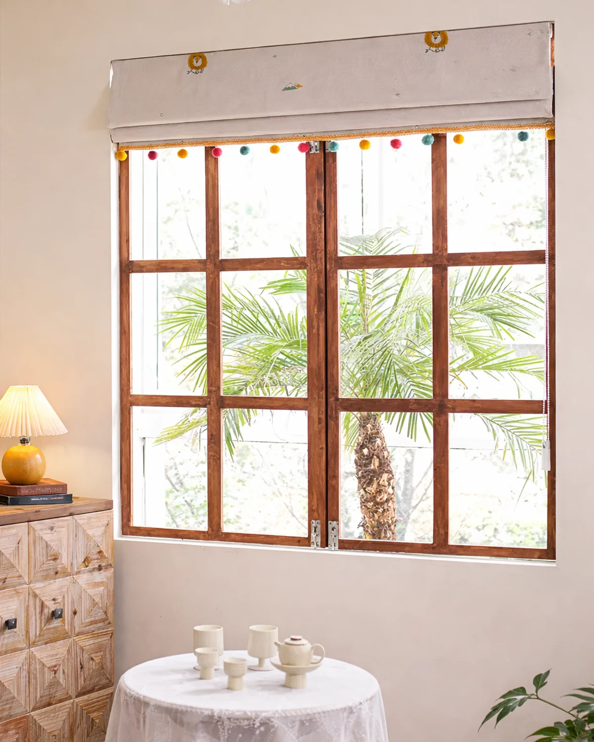 Safari embroidered Roman shade with colorful pom-pom trim, shown raised on a wooden window in a bright room.